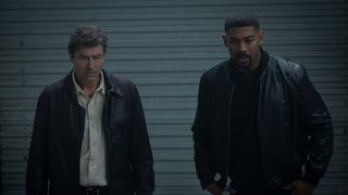 Kyle Chandler's Hal Jordan in brown jacket and shit button-up next to Aaron Pierre's John Stewart wearing black leather jacket, standing inside storage unit looking at Hal's secret safe in Lanterns.