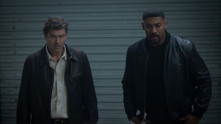 Kyle Chandler's Hal Jordan in brown jacket and shit button-up next to Aaron Pierre's John Stewart wearing black leather jacket, standing inside storage unit looking at Hal's secret safe in Lanterns.