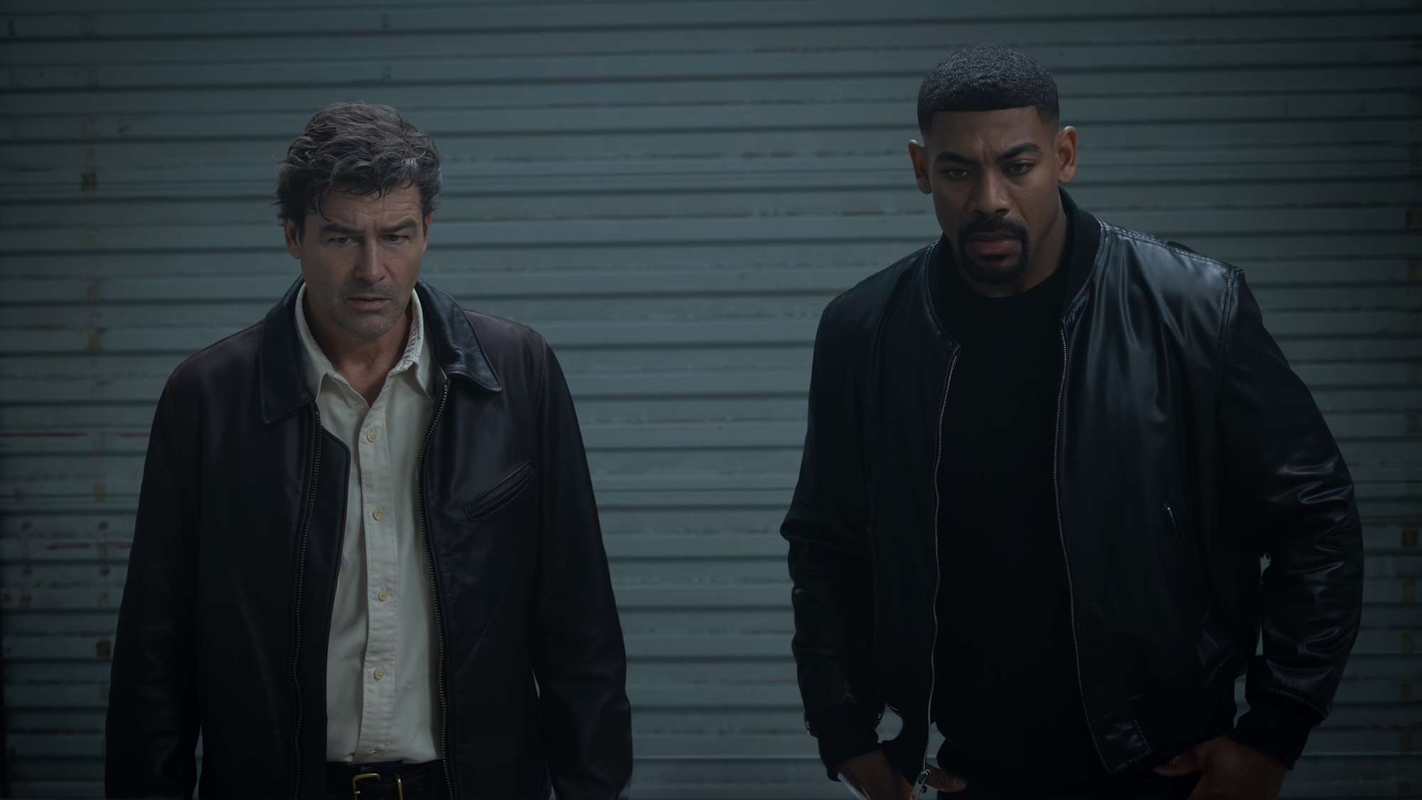 Kyle Chandler's Hal Jordan in brown jacket and shit button-up next to Aaron Pierre's John Stewart wearing black leather jacket, standing inside storage unit looking at Hal's secret safe in Lanterns.