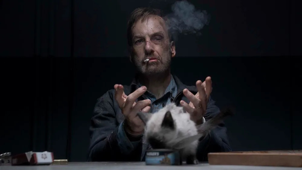 Beaten up Bob Odenkirk smoking a cigarette while handcuffed to a table while a cat eats out of a can in Nobody 2