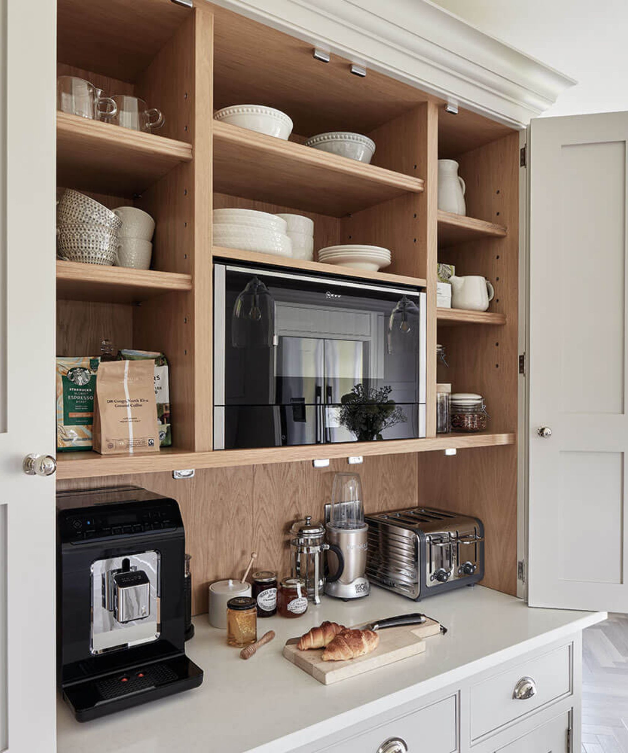 A microwave mounted into a white appliance garage with bi-folding doors. A coffee maker, blender, and toaster below, and dinnerware on the shelving above and either side.