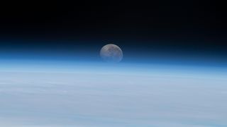 view of the full moon from the space station, showing it hanging in the blackness of space over earth's wispy blue atmosphere