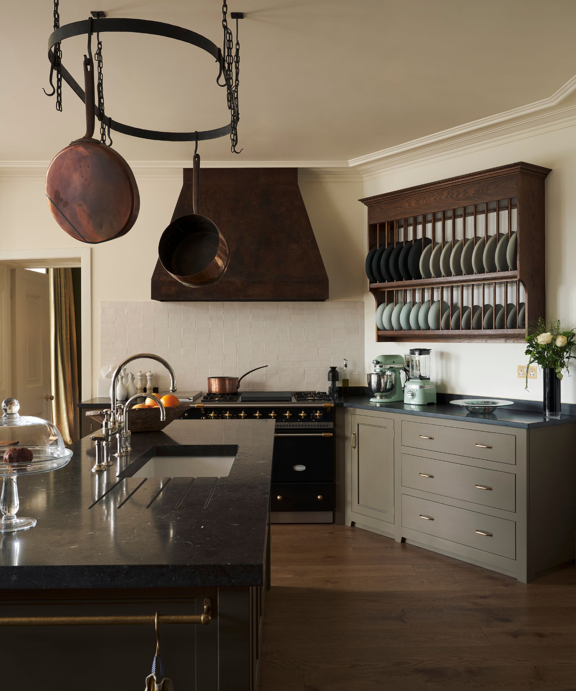Spacious kitchen with a black stone island, pale brown cabinets, hanging copper pots on a circular rack, and a dark range hood with a wooden plate rack