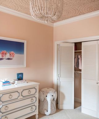 Pink children's room, wallpaper ceiling, pink painted walls, built in wardrobe with white wooden doors, white chest of drawers, blue and pink artwork, cream carpet, unique white elephant stool