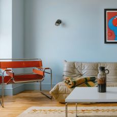 A living room painted in a baby blue shade with a contrasting red chair and artwork and a Togo sofa in beige leather by Ligne Roset