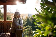 An older, attractive woman smiles and leans on a porch railing. She has a warm cup of tea or coffee and the sun is rising or setting. She exudes calm.