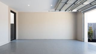 A clean, modern garage with smooth, speckled grey flooring and cream walls. The ceiling is white with recessed spotlights, and the metal garage door is partially open. A black-framed glass door leads to another part of the home, and a small tree is visible just outside next to a timber fence. 