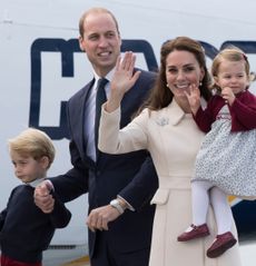 Kate Middleton waving and smiling holding a baby Princess Charlotte standing next to Prince William, wearing a blue suit and holding hands with toddler Prince George