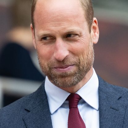 Prince William smiling while wearing a dark gray suit, a dark red tie, and a white shirt