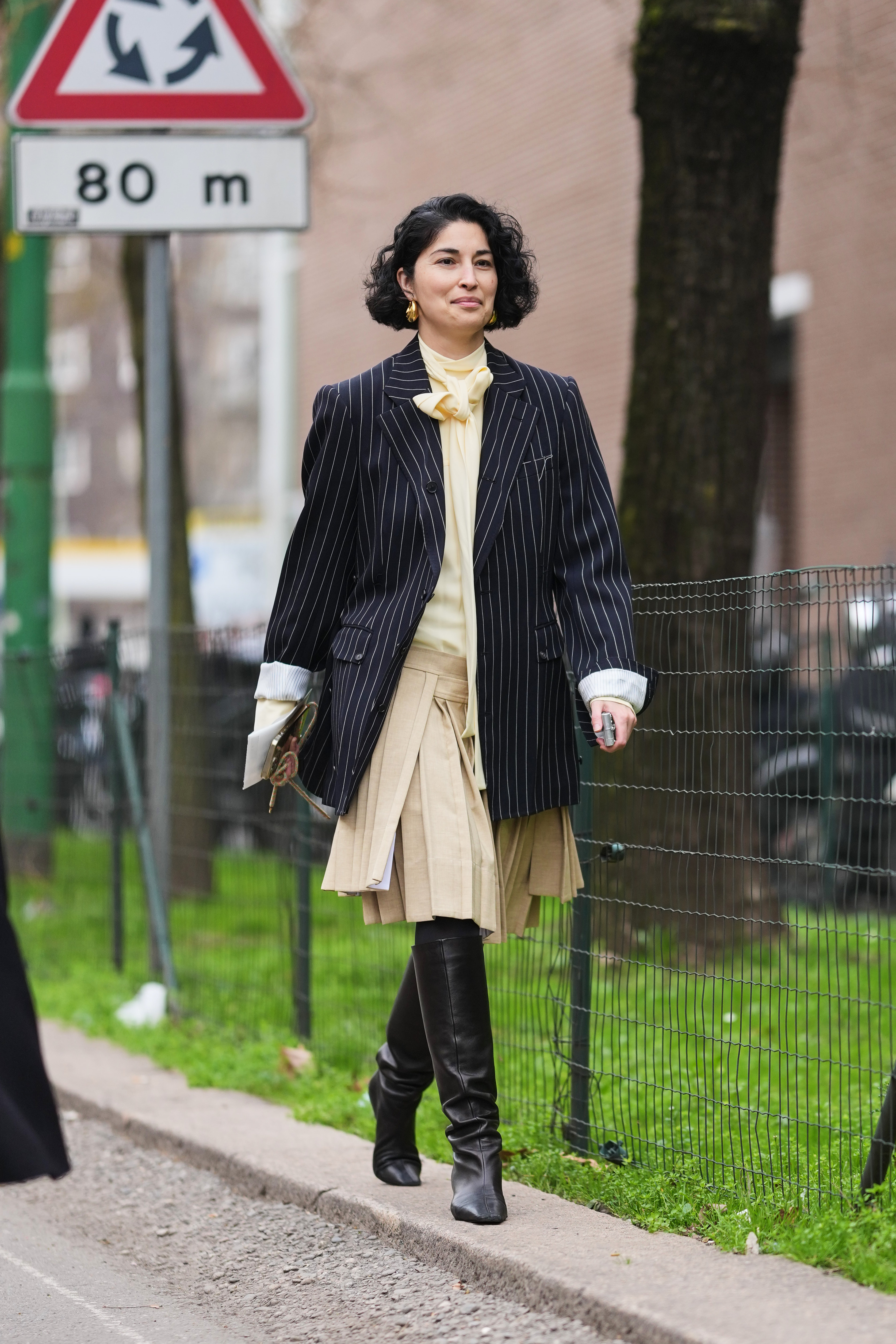 a woman wearing a pinstripe blazer, beige pleated kilt, and black knee high boots
