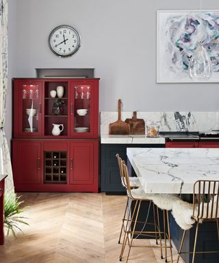 A kitchen with a burgundy red kitchen cabinet, a gray wall, marble splashback and kitchen island, gold chairs with white seats, and wooden herringbone flooring