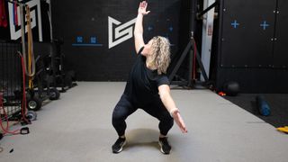 Woman in a squat with knees bent, reaches one hand up behind her. Her head is turned to look at the hand above and behind her. Her other arm is extended in front of her. She wears a black T-shirt, black leggings and black trainers.