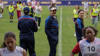 From left to right: Brendan Hunt not facing the camera, while Jason Sudeikis and Tanya Reynolds look backward and toward the camera in Ted Lasso. Women are running around them.