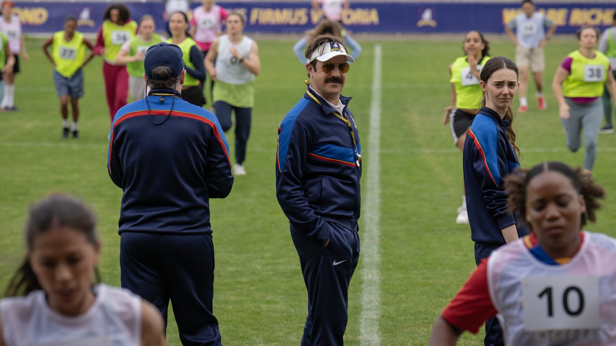 From left to right: Brendan Hunt not facing the camera, while Jason Sudeikis and Tanya Reynolds look backward and toward the camera in Ted Lasso. Women are running around them.