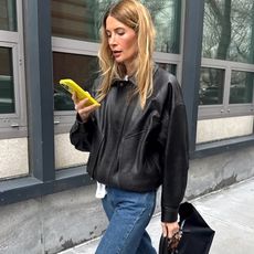 NYC style influencer Courtney Grow walks on the street while looking at her yellow phone wearing a black leather bomber jacket, jeans, and a black top-handle tote bag
