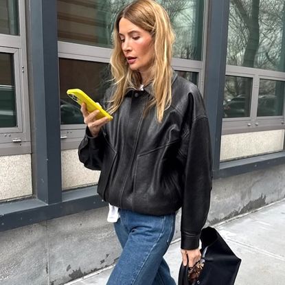 NYC style influencer Courtney Grow walks on the street while looking at her yellow phone wearing a black leather bomber jacket, jeans, and a black top-handle tote bag