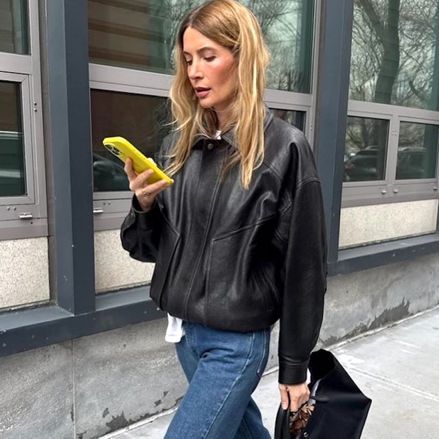 NYC style influencer Courtney Grow walks on the street while looking at her yellow phone wearing a black leather bomber jacket, jeans, and a black top-handle tote bag