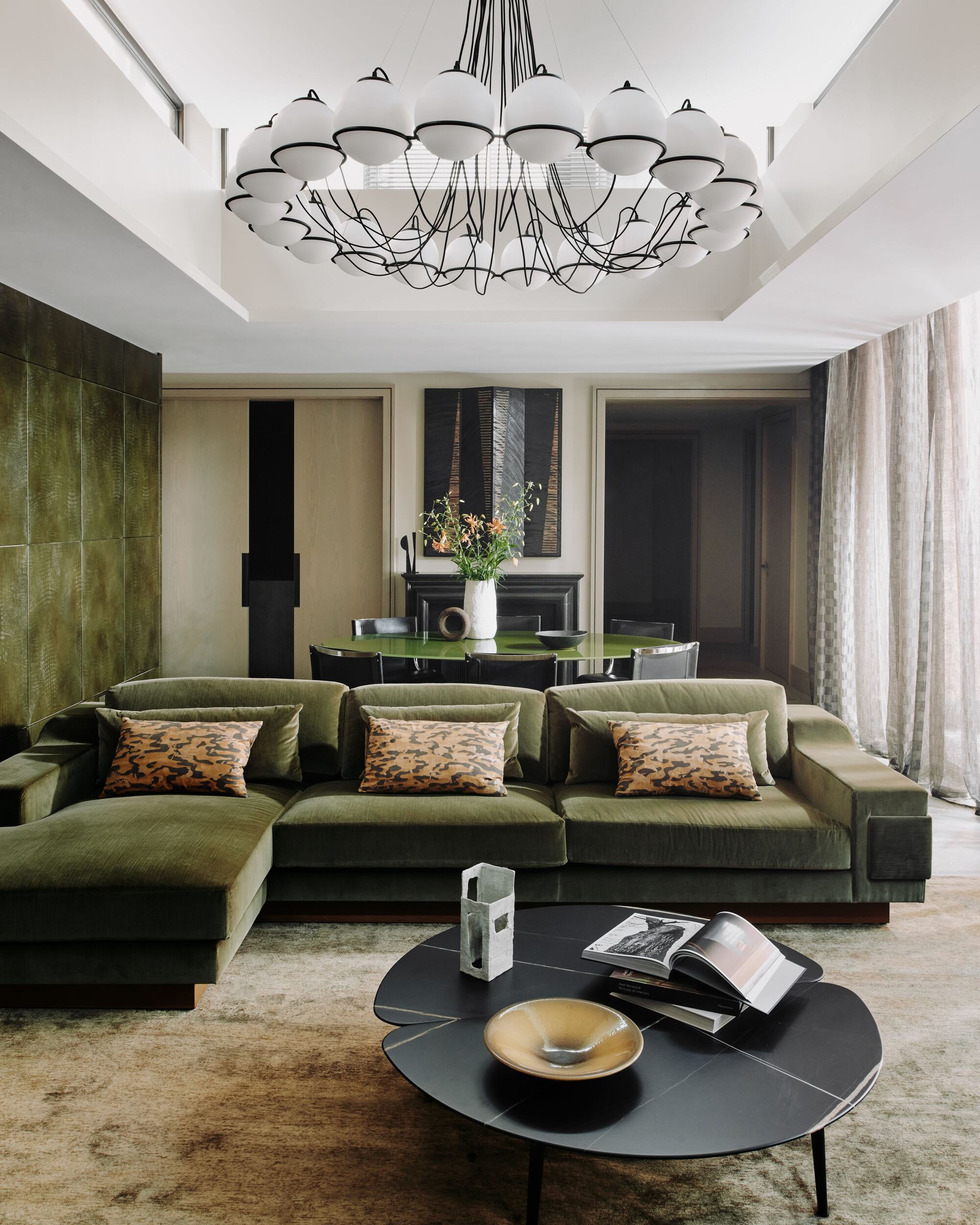 green living room with green feature wall, green velvet sectional, black round coffee table styled with bowl and books, white ceiling with statement pendant light, and checkered sheers