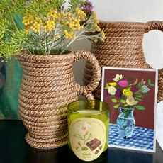 Two woven pitchers, once with fresh flowers, sitting behind floral business card and candle in green glass votive