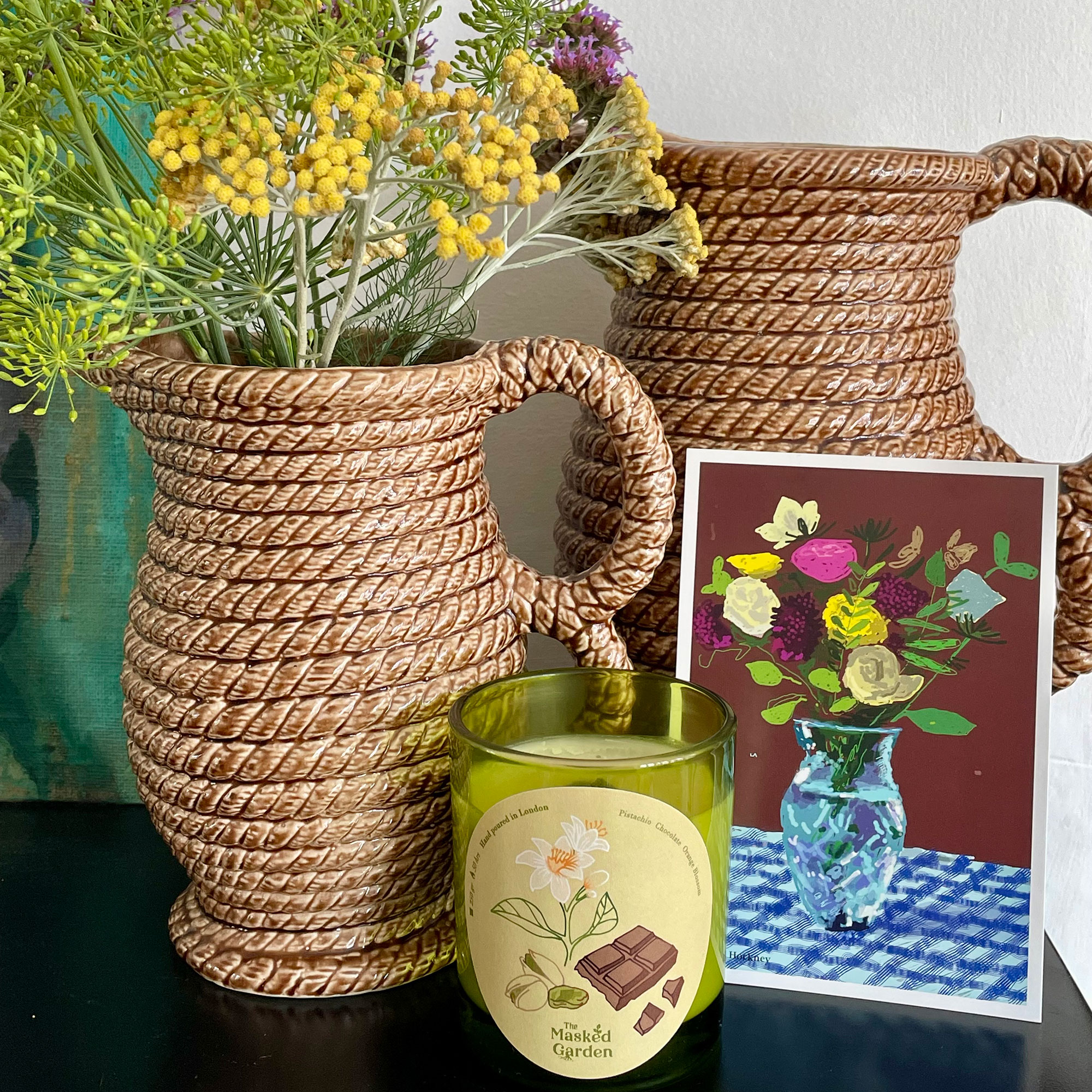 Two woven pitchers, once with fresh flowers, sitting behind floral business card and candle in green glass votive