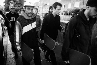 Black and white daytime, outside image of the Palace Skate Team, 2016, carrying their skateboards, surrounding buildings and hedges to the right of the shot