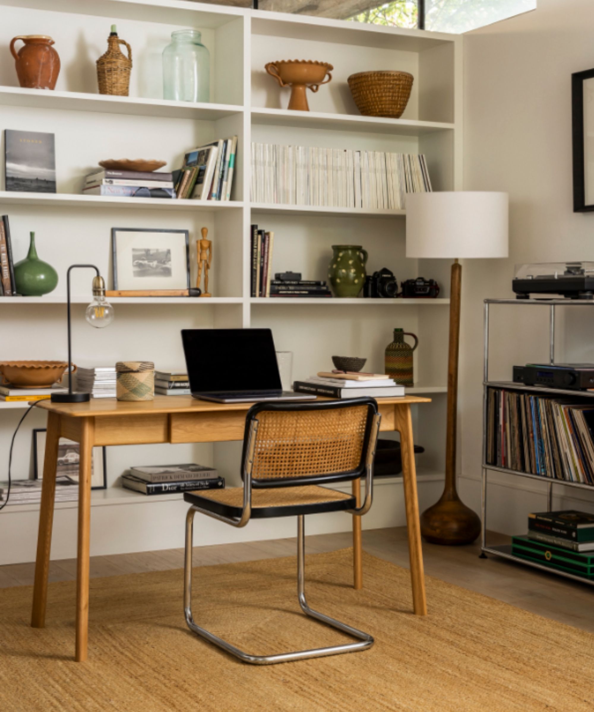 Warm mid-century home office featuring a wood desk, woven-back chair, open shelving with books and ceramics, and a record player on a metal stand.