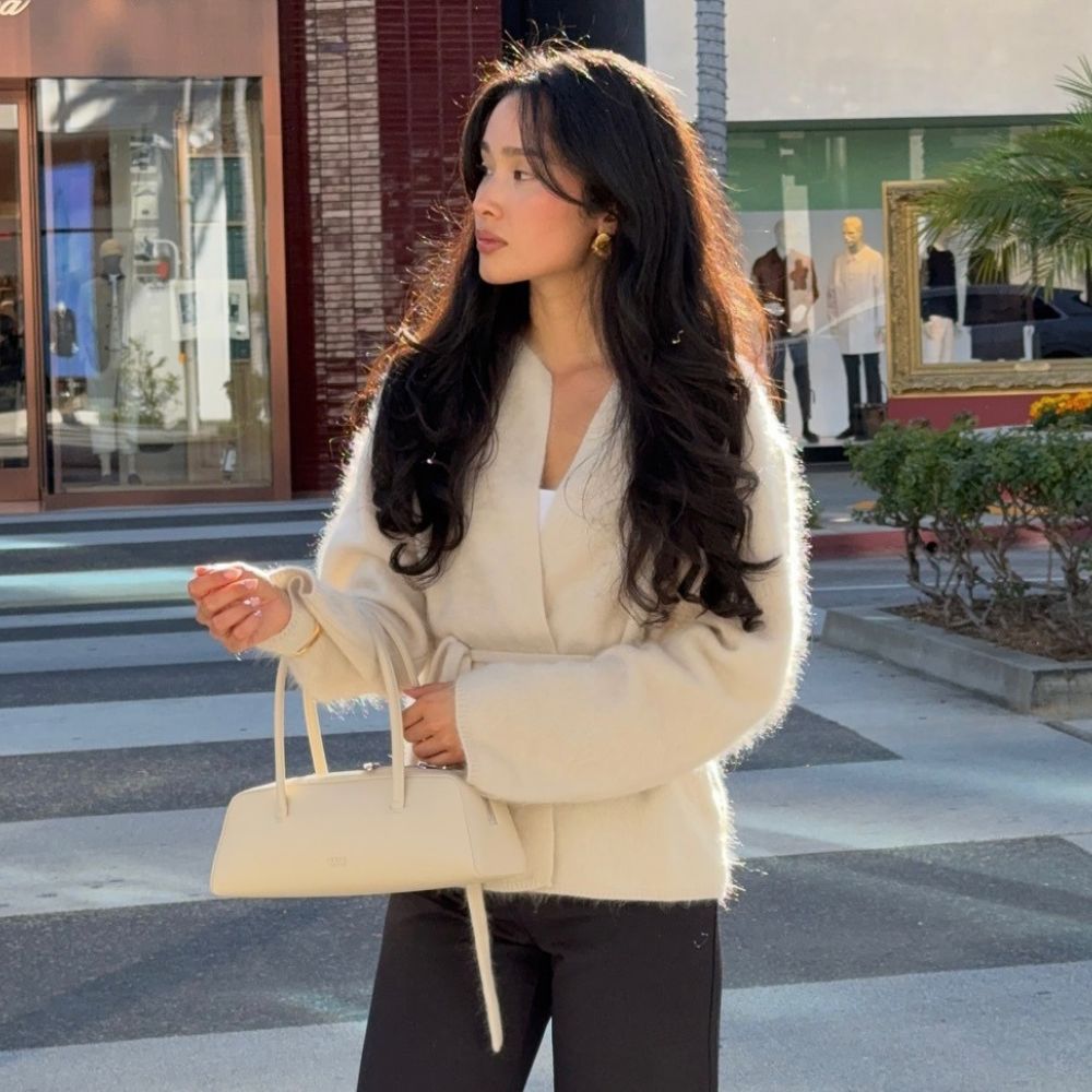 Woman wears cream brushed cardigan, carries cream bag and wears black tailored trousers