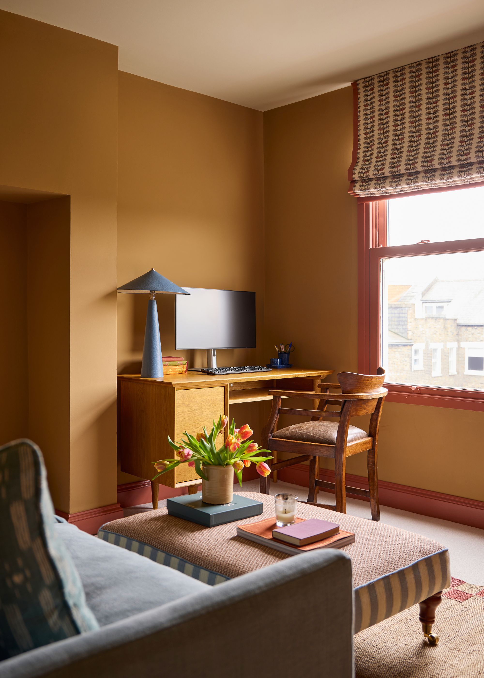 A home office room with ochre walls, red trim on the window and skirting boards, a desk with a wooden chair and a modern blue lamp, and a striped ottoman in the middle of the room.