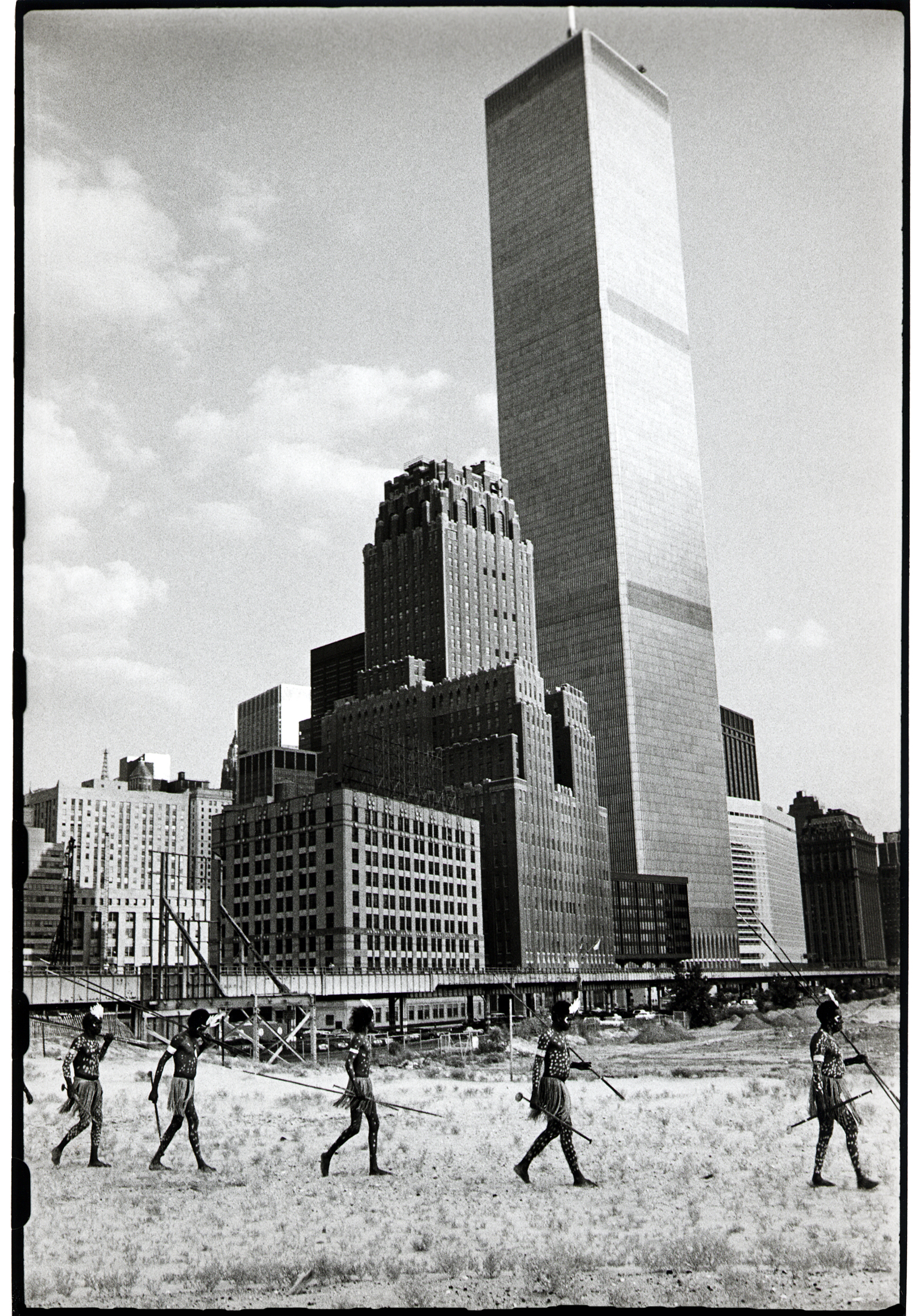 A line of Aboriginal men in body paint and grass skirts walks across wasteland carrying spears, with one of the World Trade Center towers rising behind them.&amp;rdquo;