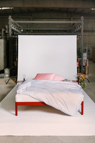 The ReFramed bed with red frame and side tables, with white bedding and pink pillows, photographed on a set in the factory. The edges of a white paper background are visible, and on the bed sides are a small lamp and a vase with flowers