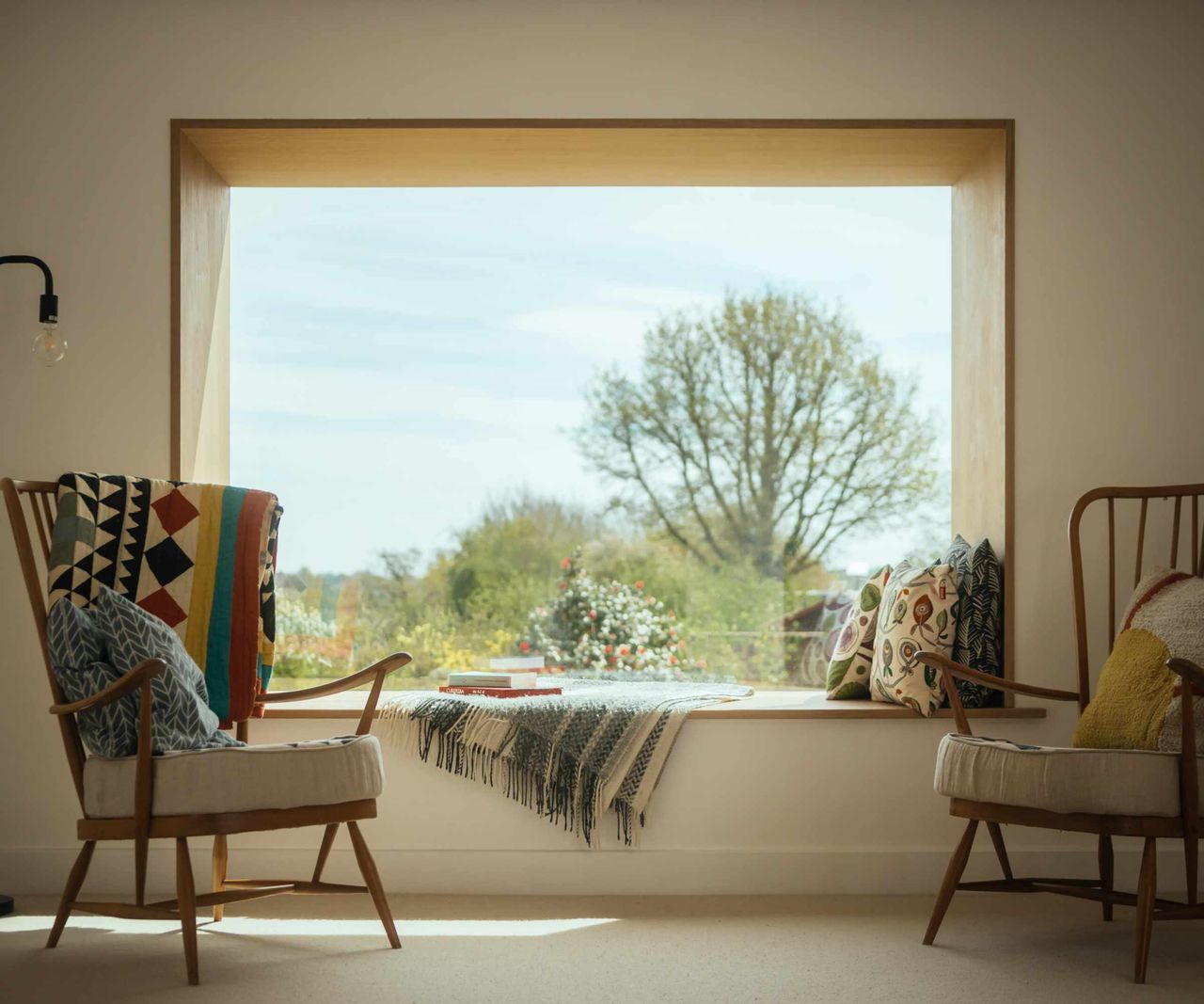 Large picture window, offering a view of trees outside, is lined with timber and features a window seat. Two Scandinavian are placed either side of the window.
