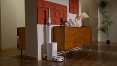 A white Switchbot k10 pro combo in a 70s style room with wooden flooring, a wooden fresser with white lamp, orange abstract wall art and a large green tree in the background. The room has white walls, and an additional room is seen beyond to the right.
