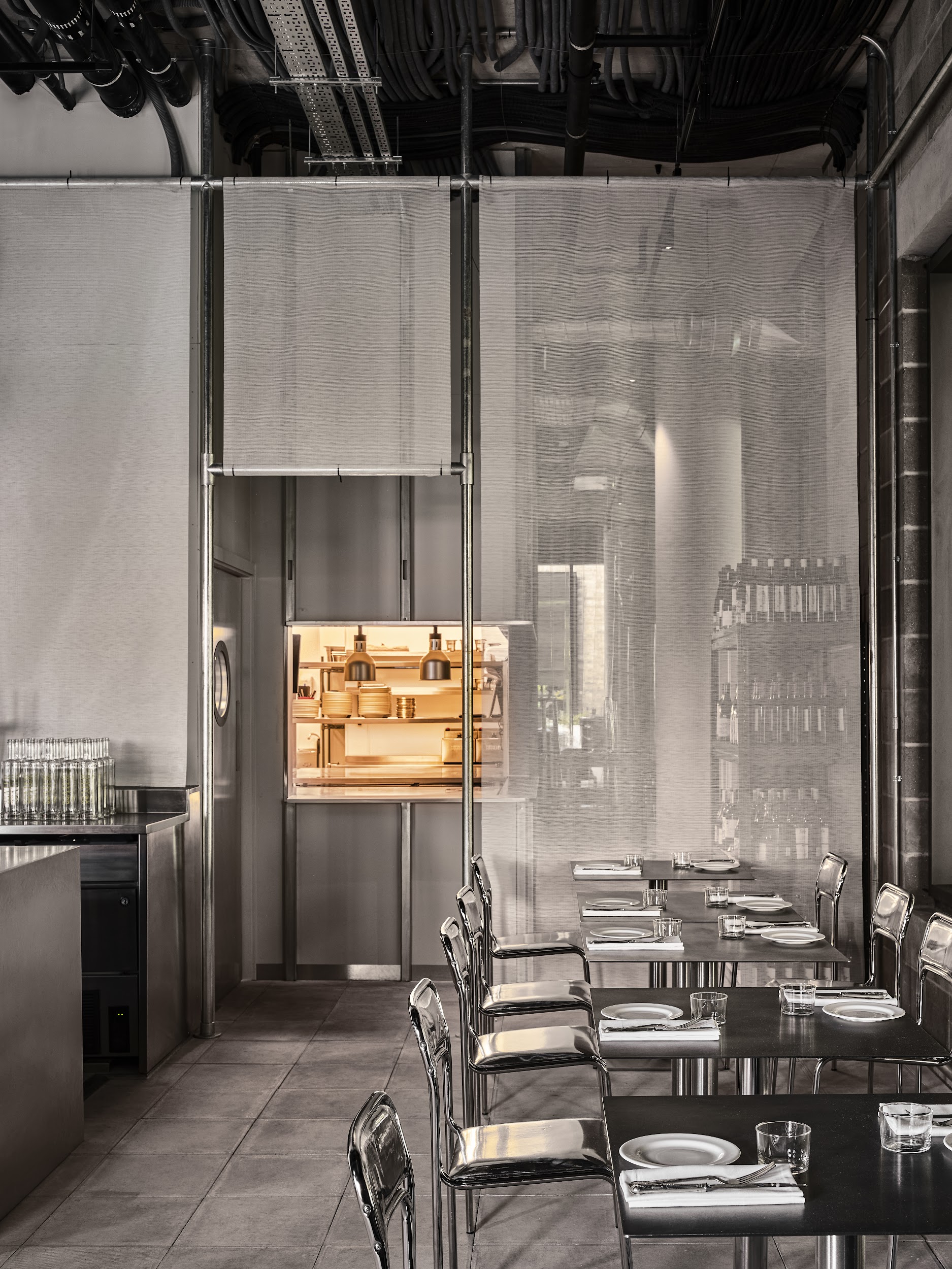 A restaurant's kitchen is concealed by semi transparent, white curtains, and releases golden-tinted warmth into an otherwise monochromatic, metallic dining room.