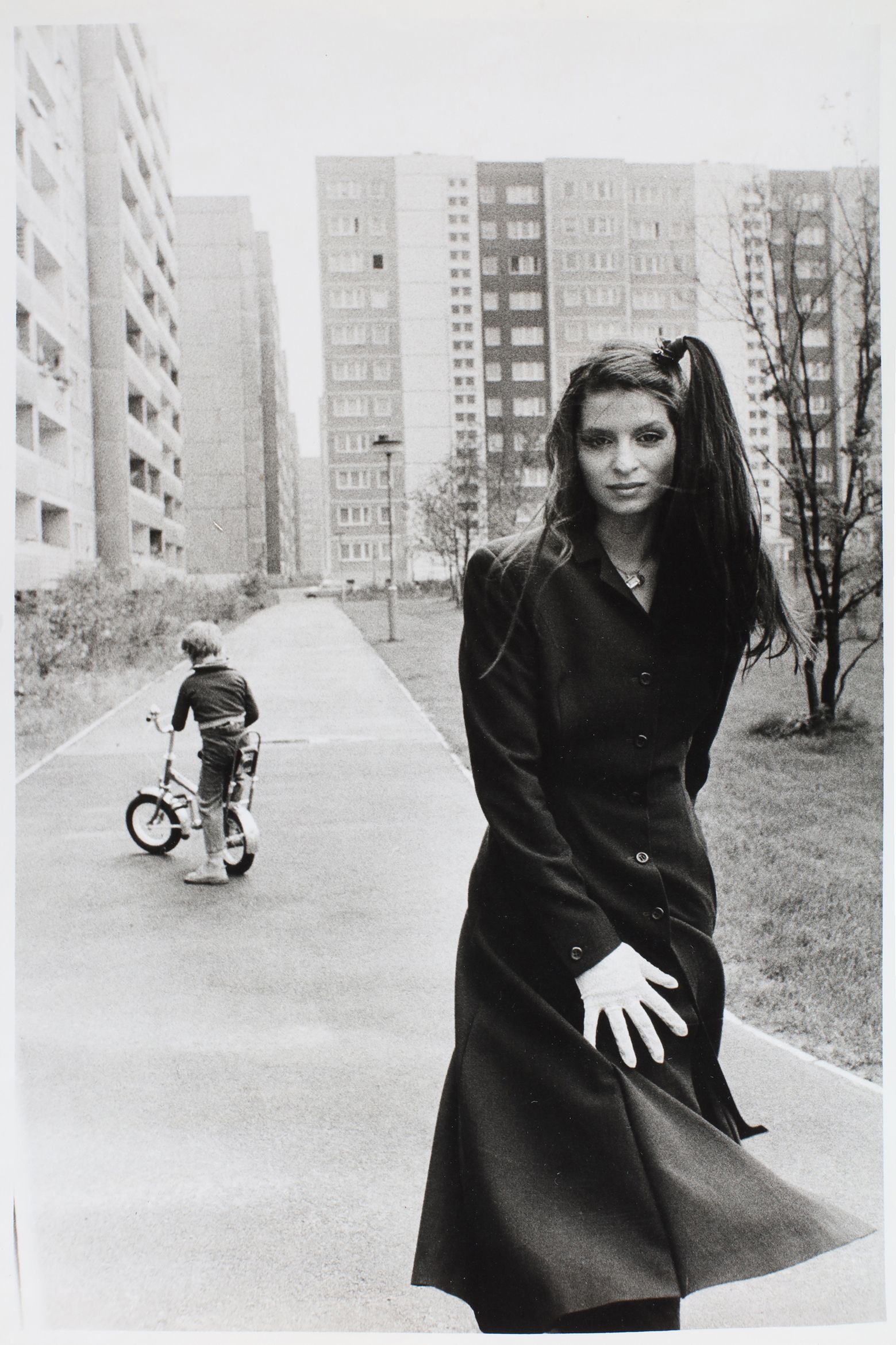 Black-and-white photo of a woman in a long coat with a child pushing a bike in an urban residential area