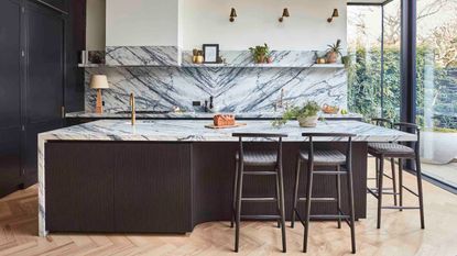 kitchen with marble backsplash and countertop, and black fluted finish with black bar stools, white walls, parquetry floors and floor-to-ceiling doors leading to the garden