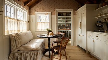 A small kitchen with white cabinets, brass hardware, and a dinette featuring a striped armless love seat, a round pedestal table, and two wooden chairs