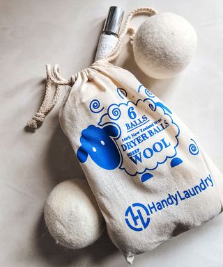 A beige bag with a blue sheep illustration on it, with two beige wool dryer balls around it and an essential oil bottle at the top of it, lying on a white surface