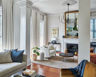 Neutral living room with sofa and armchairs, coffee tables, fireplace with mirror above, ceiling rose and chandelier and pillars with ornate plasterwork