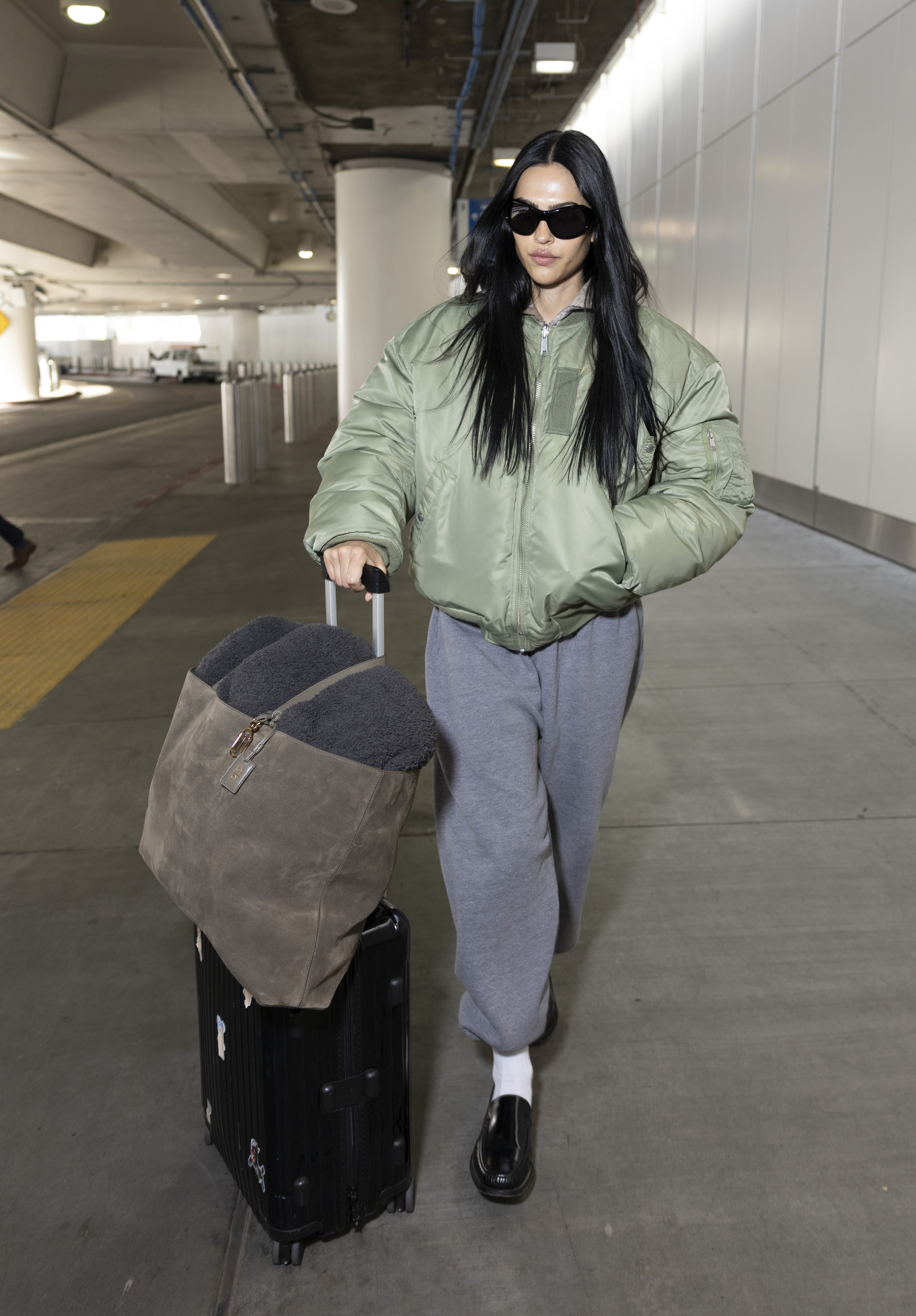 Amelia wears a green puffer jacket, gray sweatpants, white socks, black loafers, and a suede alo tote bag.