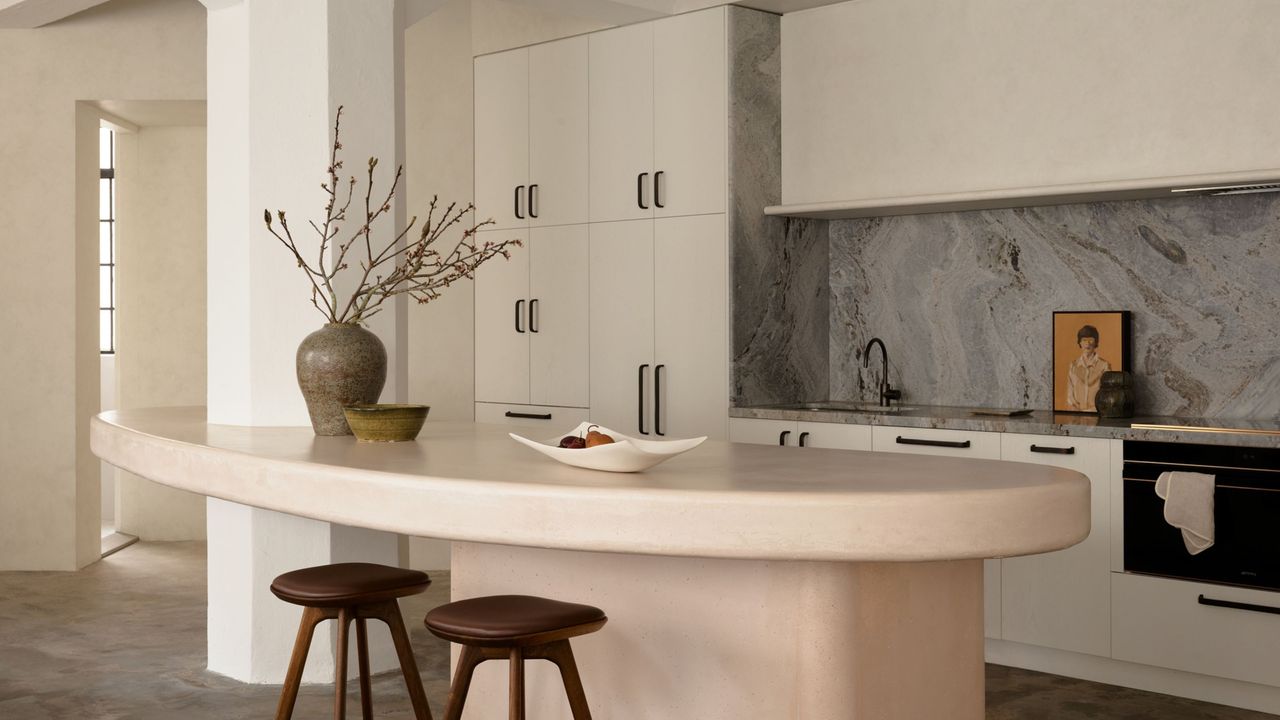 A neutral kitchen with a curved rendered blush pink kitchen island, two dark timber bar stools, a gray marble backsplash, white cabinetry with black hardware