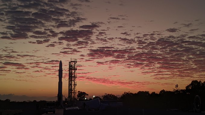 the silhouette of a rocket stands against a soft orange sky at sunset.