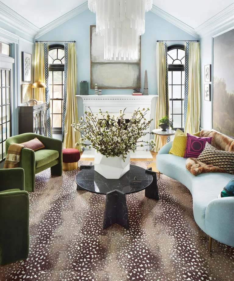 Colorful blue living room with blue curved sofa, two green armchairs, fireplace, yellow curtains, chandelier, animal print brown rug, black coffee table with vase of flowers