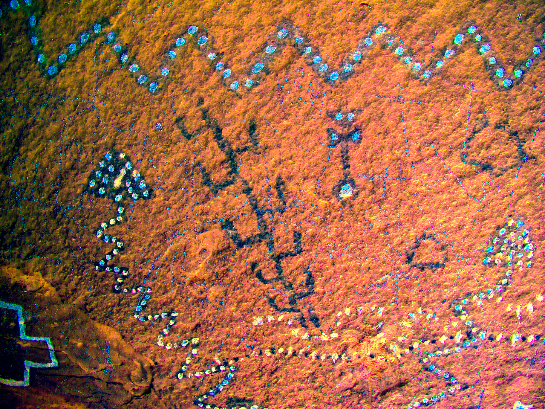 A close-up rock panel processed with decorrelation stretch in teal and orange tones, revealing zigzag lines, dot clusters and animal-like figures invisible in the original photograph.