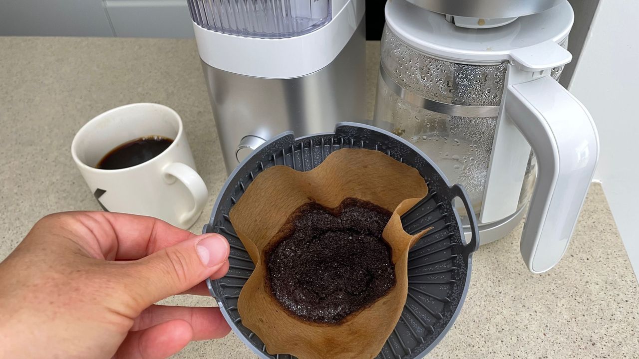 A close up shot of wet coffee grounds in a paper filter, with a drip coffee maker blurry in the background.