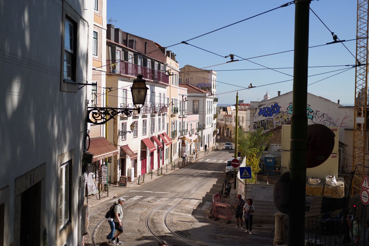 Sunlit Lisbon street with tram tracks, pastel buildings, overhead wires and pedestrians crossing near a construction site.