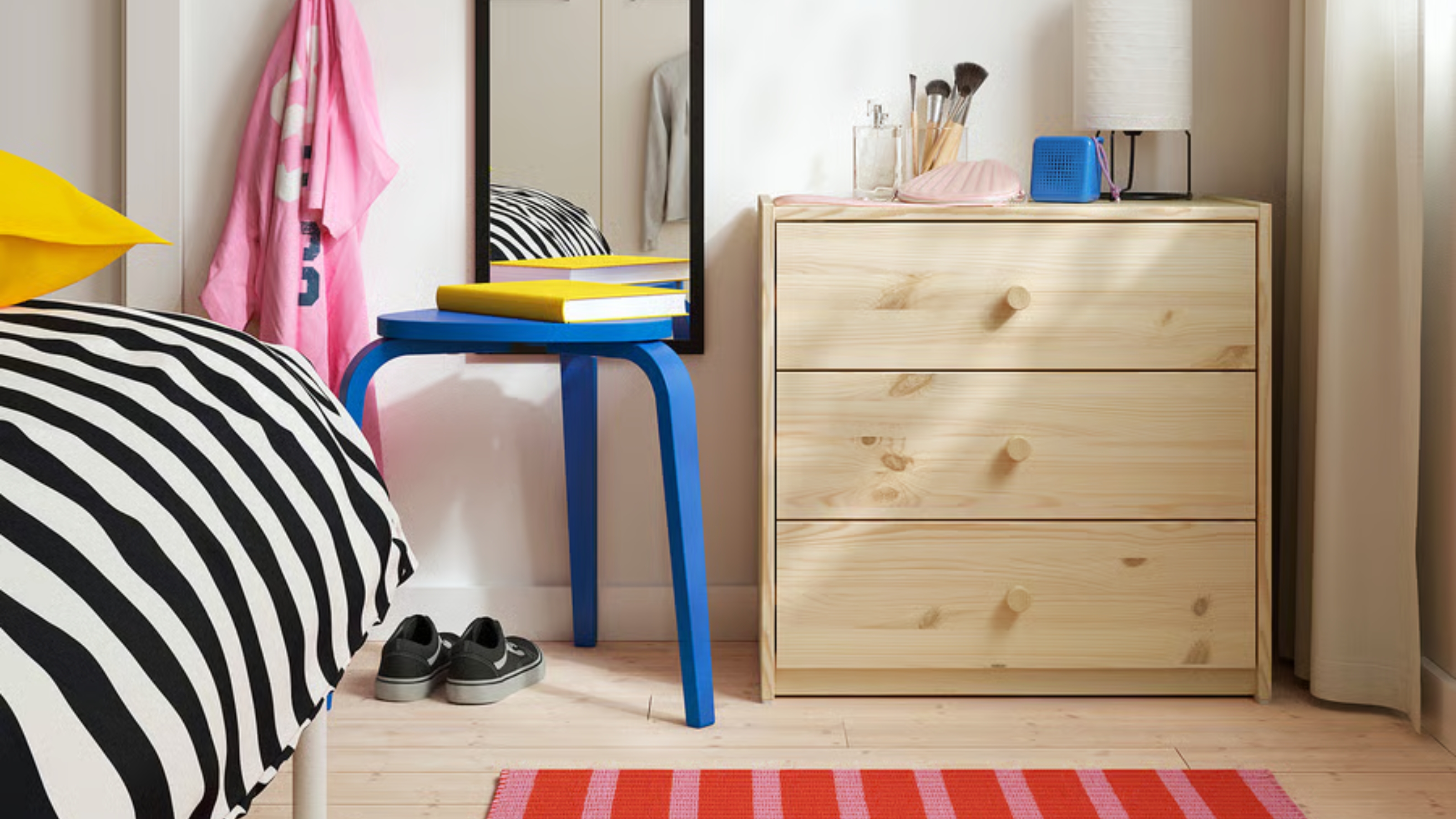 Image of a white bedroom wall with a plain wooden chest of three drawers. There is an electric blue stool and a bed that has black and white bedding.
