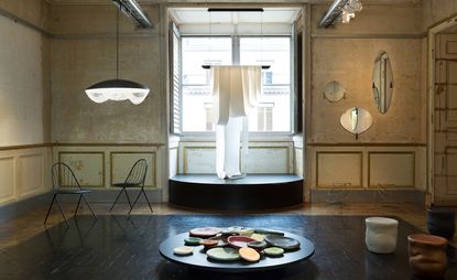 A room with run-down walls with mirrors hanging to the right, a big window in the center, with an art piece hanging from the ceiling. Low table with ceramic plates in different sizes and colors, and two black chairs to the left.