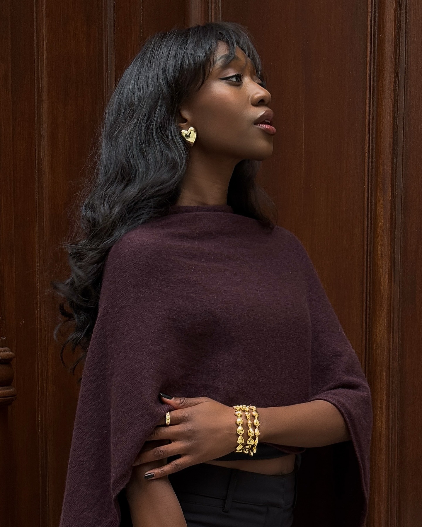 Close shot of Influencer @emmanuellek_ wearing a dark aubergine cape knit with gold earrings and a gold bracelet and ring.
