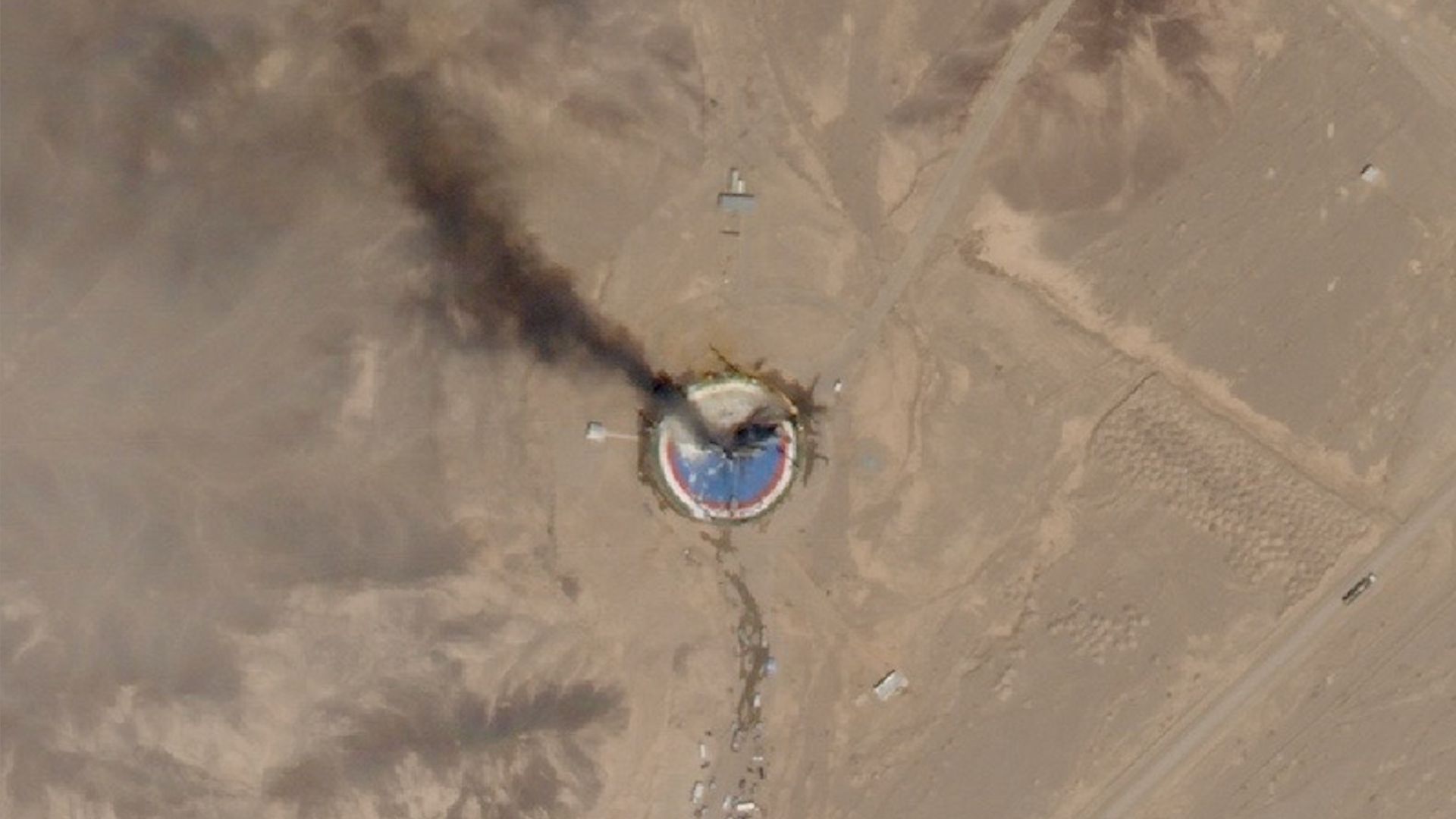 satellite photo of a circular, blue launch pad in the desert with black smoke rising from it
