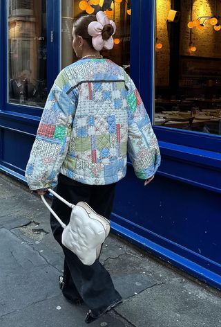 quilted patchwork jacket trend is shown on a woman walking down the street wearing a floral scrunchie, cropped coat, jeans, ballet flats, and a white shoulder bag